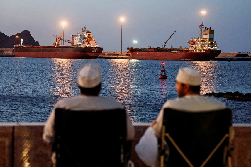 Two men watching oil tankers anchored at dusk as ships wait outside the Strait of Hormuz amid the closure